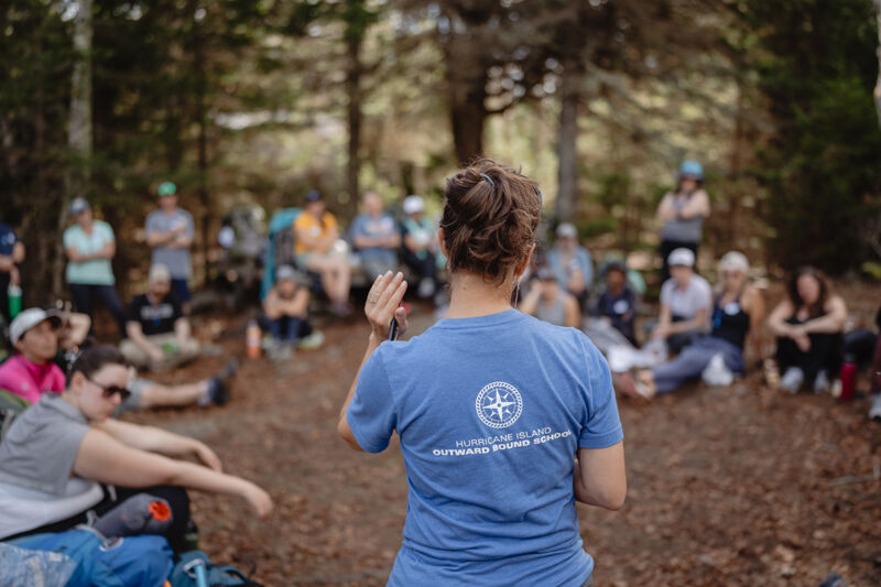 The image shows a group of people gathered in a forest. A woman in a blue shirt with a logo on the back is facing the group and appears to be addressing them. The people are sitting on the ground, and some are looking at the woman. The scene suggests a meeting or outdoor activity.